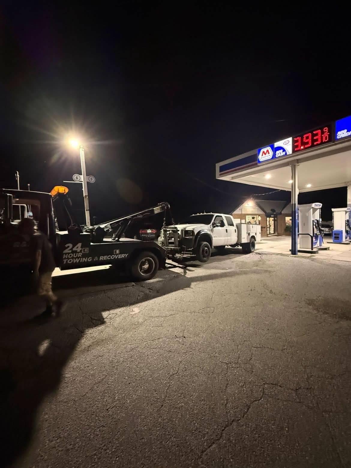 CARSTAR wheel lift tow truck recovering a white utility truck at a gas station at night