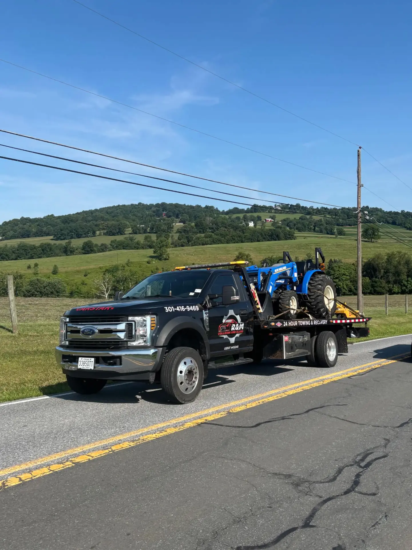 CARSTAR flatbed tow truck hauling a blue tractor on a rural Maryland road